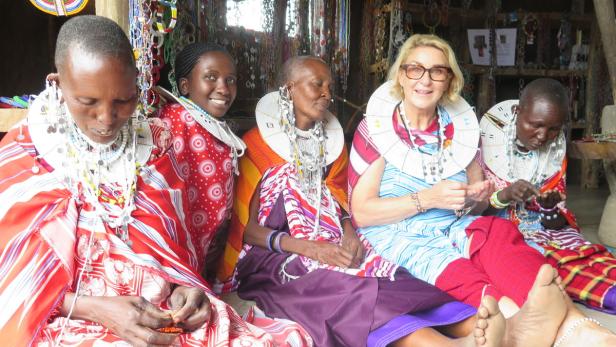 Eine westliche Frau sitzt mit vier Massai-Frauen zusammen, die traditionelle Kleidung und Schmuck tragen.