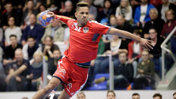 Ein Handballspieler im roten Trikot wirft den Ball vor einem Publikum.