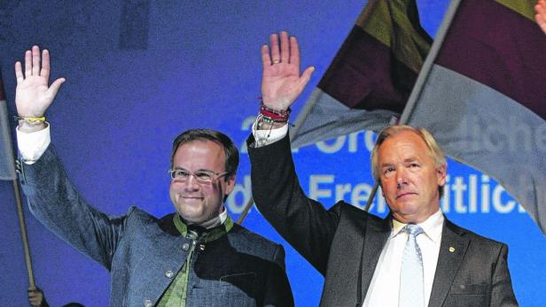 Zwei Männer winken vor einem blauen Hintergrund mit bayerischen Flaggen.