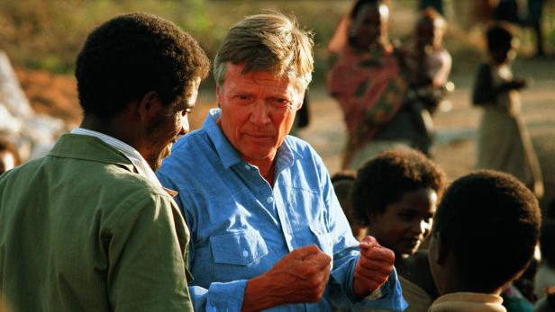 Ein Mann in einem blauen Hemd spricht mit einer Gruppe von Menschen im Freien.