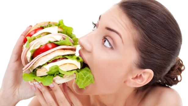 Young woman biting into a very big homemade bread roll filled with tomato and bacon on a white background