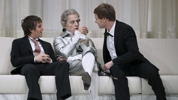 Drei Männer sitzen auf einem Sofa, einer davon in historischer Kleidung.