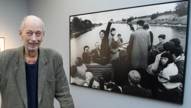 Ein Mann steht vor einem Schwarzweißfoto einer Gruppe von Menschen auf einem Boot.
