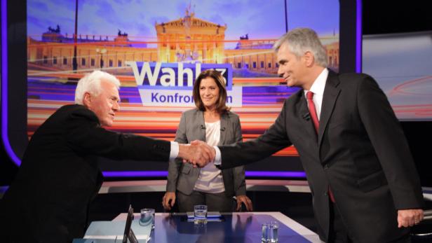 Zwei Männer schütteln sich vor einer „Wahl 13“-Kulisse die Hände, während eine Frau zusieht.