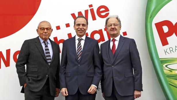 Drei Männer in Anzügen posieren vor einem Persil-Werbeplakat.