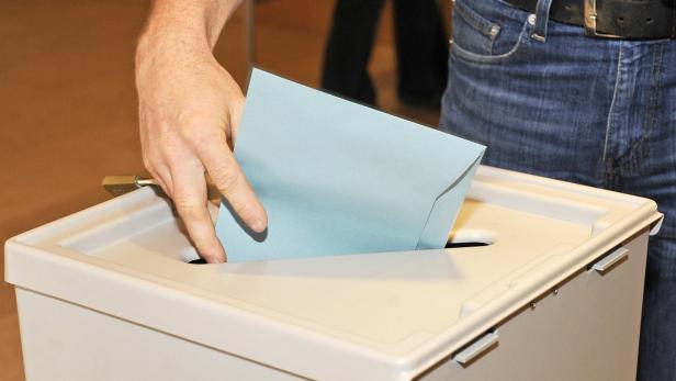 Eine Person wirft einen blauen Briefumschlag in eine Wahlurne.