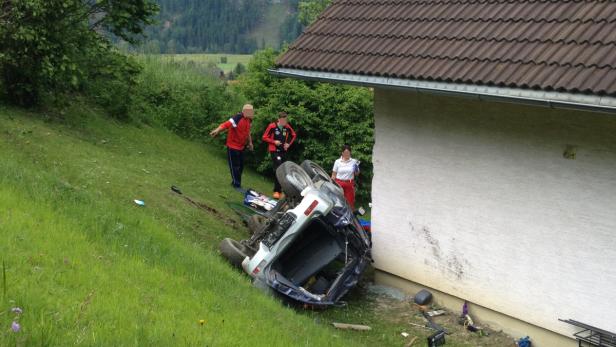 Ein Auto ist einen Hang hinuntergerollt und gegen eine Hauswand geprallt; drei Personen stehen daneben.