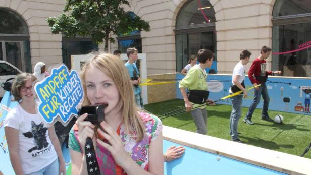 Ein Mädchen bläst in eine Trillerpfeife, während im Hintergrund Kinder Fußball spielen und ein Schild „Andiff für Kinder Rechte!“ zu sehen ist.