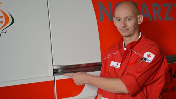 Ein Sanitäter in roter Uniform steht vor einem Rettungswagen und zeigt auf die Tür.
