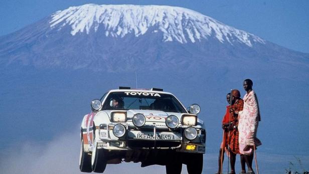 Ein Toyota-Rallyeauto springt vor dem Kilimandscharo, neben dem zwei Massai stehen.