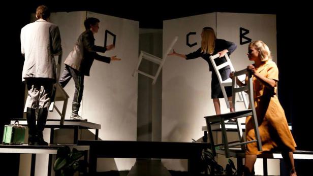 Eine Theaterszene mit vier Schauspielern und fliegenden Stühlen vor weißen Wänden mit Buchstaben.