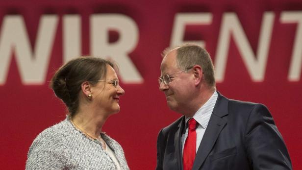 Zwei Personen, eine Frau und ein Mann, stehen vor einem roten Hintergrund mit weißer Schrift.