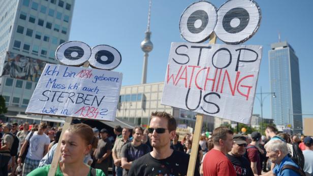 Menschen demonstrieren mit Schildern, auf denen „Stop Watching Us“ und „Ich bin 1984 geboren. Muss ich auch sterben wie in 1984?“ steht.