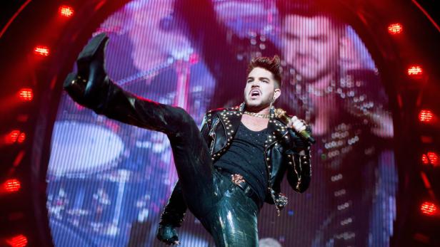 Adam Lambert tritt in glänzender Hose und Lederjacke auf einer Bühne auf.