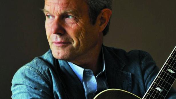 Ein Mann mittleren Alters mit grauen Haaren hält eine Gitarre im Arm.