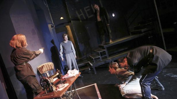 Auf einer Theaterbühne bedroht eine Frau mit einer Pistole eine andere, während ein Mann zusieht.