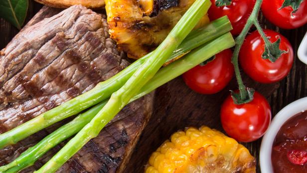 Ein Steak mit gegrilltem Mais, Spargel, Kartoffelecken und Tomaten auf einem Holzbrett.