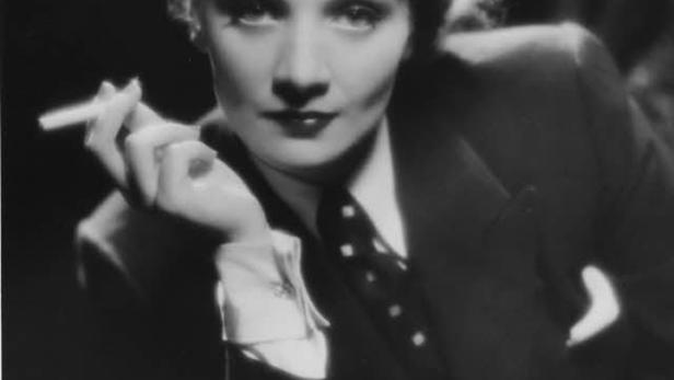 Marlene Dietrich in einem Anzug mit Krawatte, eine Zigarette haltend.