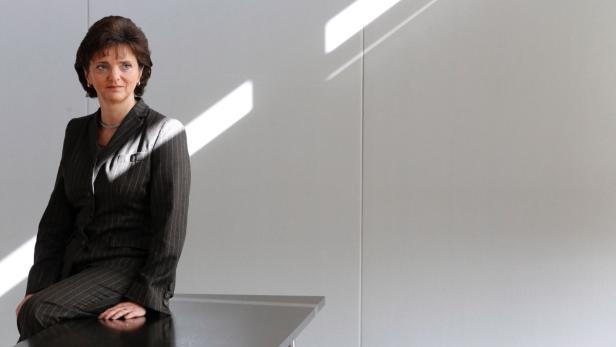 Eine Frau im Hosenanzug sitzt auf einem Tisch vor einer weißen Wand.