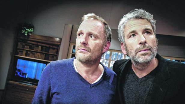 Zwei Männer blicken in einem Raum mit Bücherregal und Fernseher nach oben.