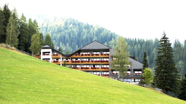 Ein Hotel mit blumengeschmückten Balkonen liegt auf einem grünen Hügel vor einem bewaldeten Berg.
