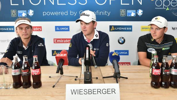 Drei Golfspieler sitzen bei einer Pressekonferenz hinter einem Tisch mit Mikrofonen und Getränken.