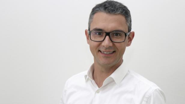 Ein lächelnder Mann mit Brille und weißem Hemd vor einem hellen Hintergrund.