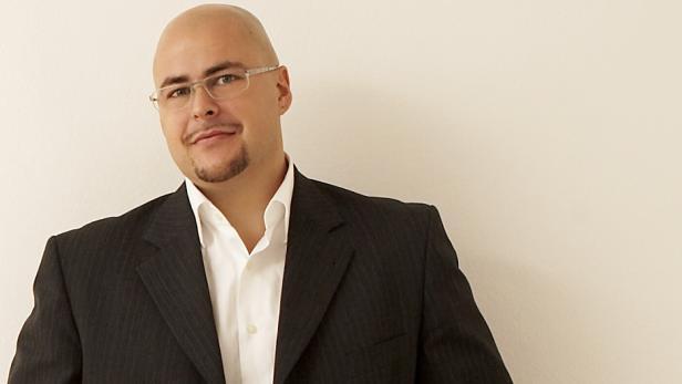Ein Mann mit Brille und Anzug vor einer hellen Wand.