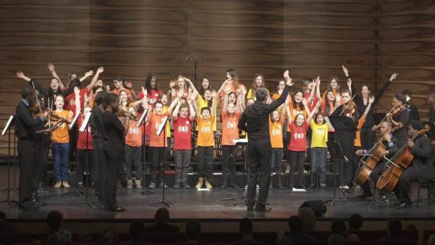 Ein Kinderchor singt auf einer Bühne, begleitet von einem Orchester.