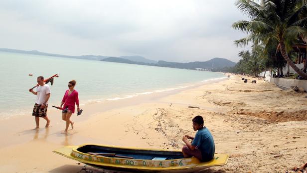 Ein Junge sitzt in einem Boot am Strand, während ein Paar im Hintergrund spaziert.
