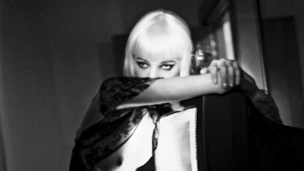 Eine Frau mit blonder Perücke lehnt an einem dunklen Möbelstück und hält ein Weinglas.