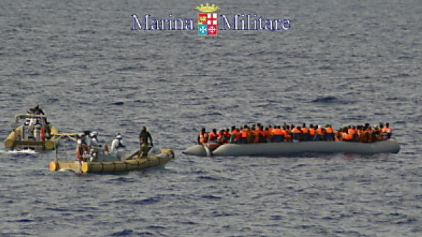 Ein überfülltes Schlauchboot mit Menschen wird von Booten der Marina Militare begleitet.