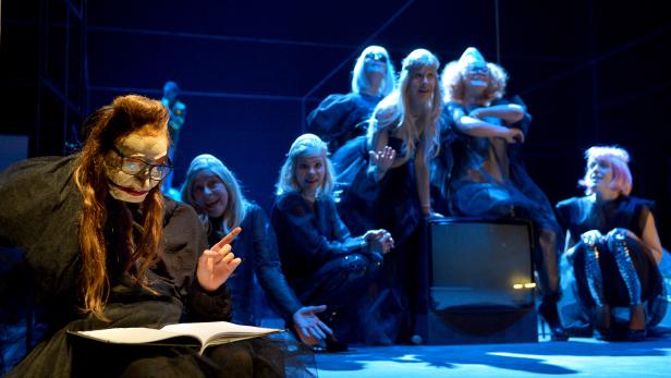 Eine Theaterszene mit einer Gruppe von Schauspielerinnen in Kostümen und Masken auf einer Bühne.