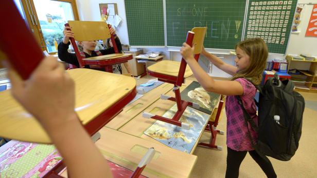 Kinder stellen Stühle auf die Tische in einem Klassenzimmer, auf der Tafel steht „Schöne Ferien!“.
