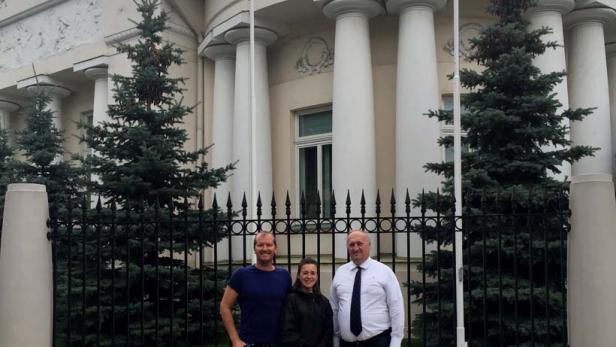 Drei Personen stehen vor einem Gebäude mit Säulen, einer lettischen und einer EU-Flagge.