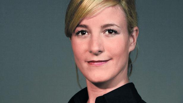 Porträt einer Frau mit blonden Haaren und einem schwarzen Hemd vor einem grauen Hintergrund.