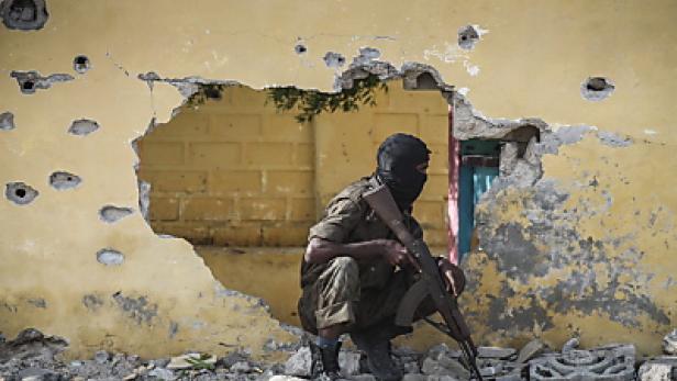 Ein maskierter Mann mit einem Gewehr kauert vor einem beschädigten Gebäude.