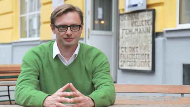 Ein Mann mit Brille und grünem Pullover sitzt an einem Tisch im Freien.
