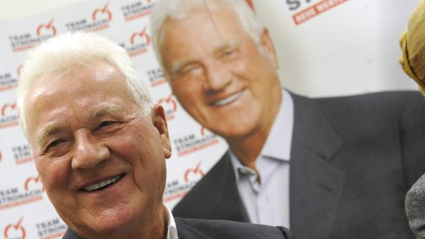 Frank Stronach lächelt vor einem Poster von sich selbst.