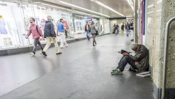 Ein Mann sitzt am Boden, während Passanten an Schaufenstern vorbeigehen.