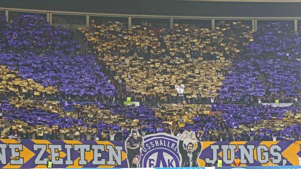 Zuschauer im Stadion halten goldene und lila Folien hoch, um ein Muster zu erzeugen.