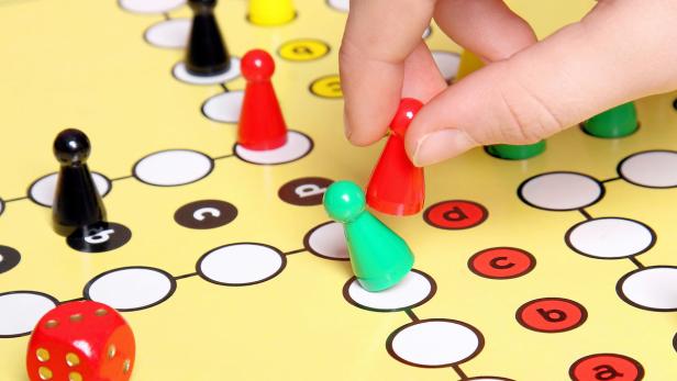 Eine Hand setzt eine rote Spielfigur auf ein gelbes Spielbrett mit bunten Figuren und einem Würfel.