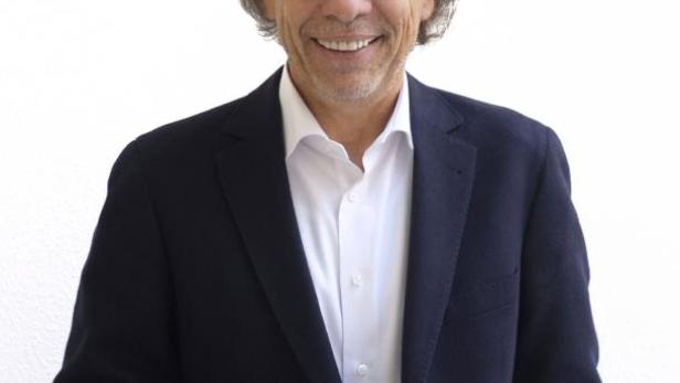 Ein lächelnder Mann mit Brille und dunklem Sakko vor einem weißen Hintergrund.