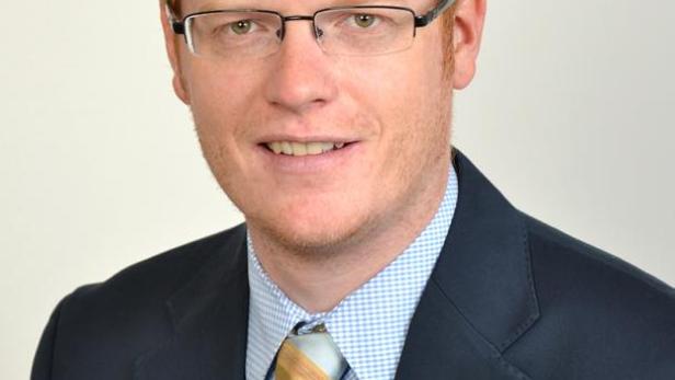 Ein Mann mit roten Haaren, Brille und Anzug blickt in die Kamera.