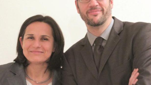 Eine Frau und ein Mann in Anzügen stehen nebeneinander und lächeln.