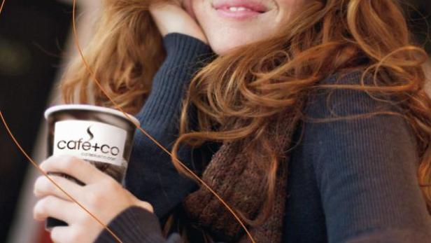 Eine lächelnde Frau mit roten Haaren hält einen Kaffee von Café+co.