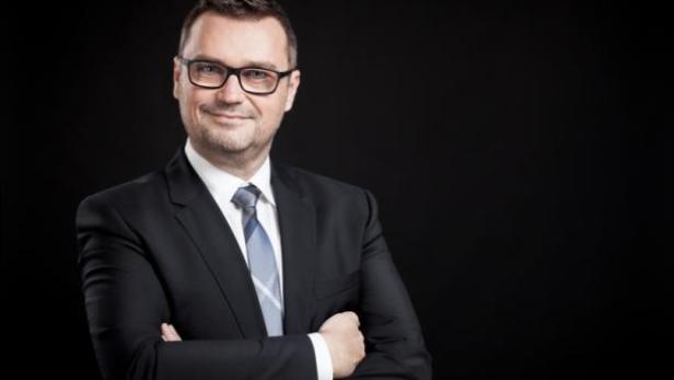 Ein Mann mit Brille und Anzug lächelt mit verschränkten Armen vor einem schwarzen Hintergrund.