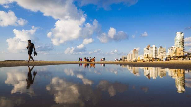 Ein Mann geht barfuß am Strand von Tel Aviv entlang, das sich im Wasser spiegelt.