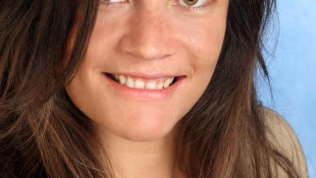 Eine lächelnde Frau mit braunen Haaren und grünen Augen vor blauem Hintergrund.