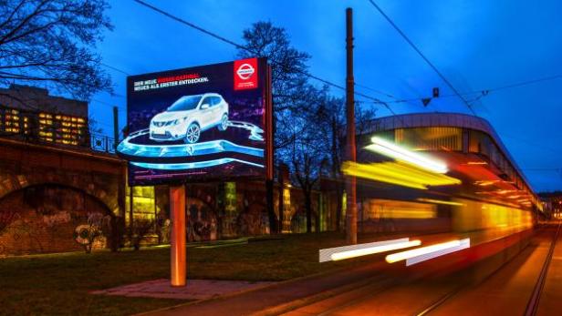 Eine beleuchtete Werbetafel für Nissan neben einer vorbeifahrenden Straßenbahn bei Nacht.